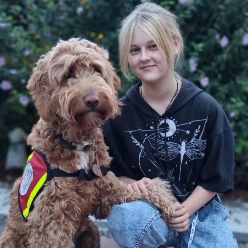 Auf dem Bild kniet eine junge Frau, links daneben sitzt ein honigbrauner gelockter Hund. Eine Pfote des Hundes liegt auf dem Knie der jungen Frau. Der Hund trägt eine rote Kenndecke.