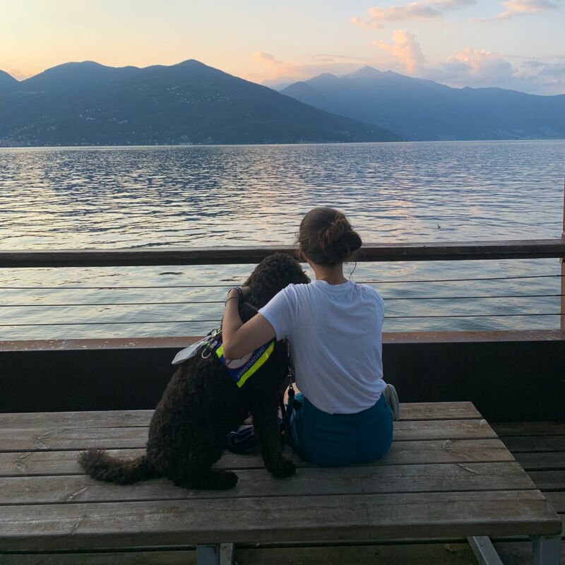 Auf dem Bild sieht man eine Frau auf einer Bank sitzen. Neben ihr sitzt ein Hund, diesen umarmt die Frau mit ihrem linken Arm. Beide sind nur von hinten. Beide schauen in Richtung des Sees und den Bergen, welche von der Bank aus zu sehen sind.