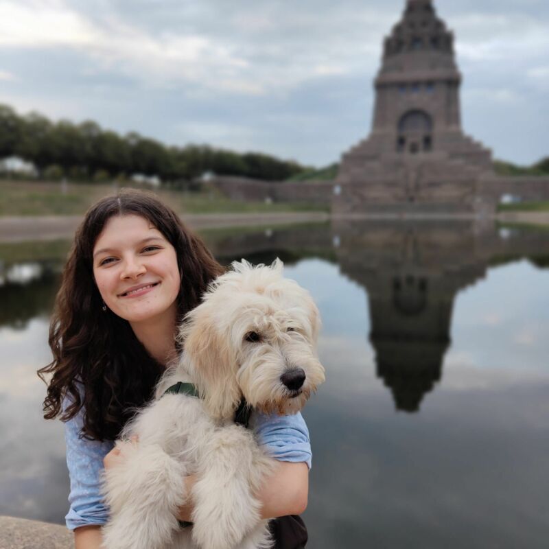 Es ist eine junge Frau zu sehen, welche auf dem Schoß einen weißen Hund sitzen hat. Die Frau lächelt in die Kamera. Die beiden sitzen vor einem großen Teich und im Hintergrund ist ein Gebäude zu sehen.